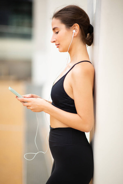 Sporthíreket viselő fiatal nő oldalsó portréja, amint edzés után egy falnak támaszkodva zenét hallgat mobiltelefonnal és fülhallgatóval - Fotó, kép