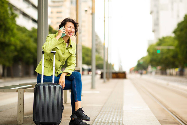 Щаслива молода жінка, спираючись на валізу і розмовляючи по мобільному телефону, чекаючи на автобусній зупинці в місті
 - Фото, зображення