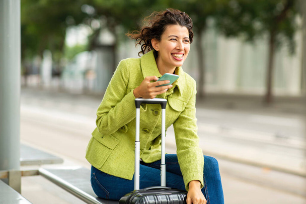 Cheerful young woman with luggage and mobile phone outdoors at city bus stop - Fotoğraf, Görsel