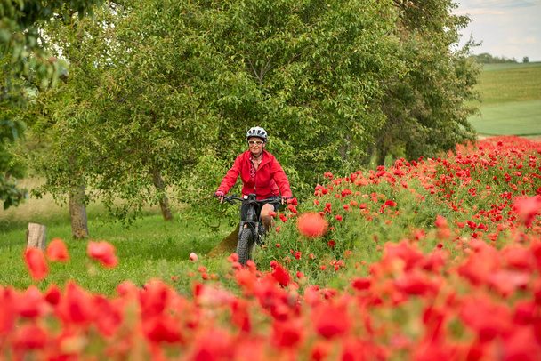 kedves, aktív idős nő szórakozás saját elektromos kerékpár egy hatalmas területen virágzó vörös mák  - Fotó, kép