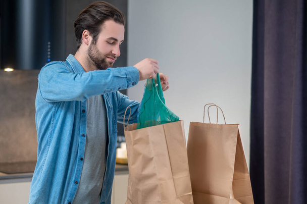 Kellemes foglalkozás. Fiatal szakállas férfi boldogan vásárol csomagokat a modern konyhában otthon - Fotó, kép