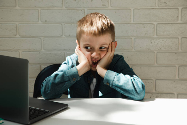 Çocuk internette öğrenmekten bıktığı için yüzünü buruşturuyor. Masadaki bir çocuğun portresi. - Fotoğraf, Görsel