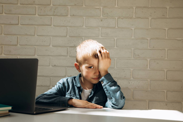 A fiú megdörzsöli a szemét az online tanulástól. Egy iskolás fiú portréja az íróasztalnál. - Fotó, kép