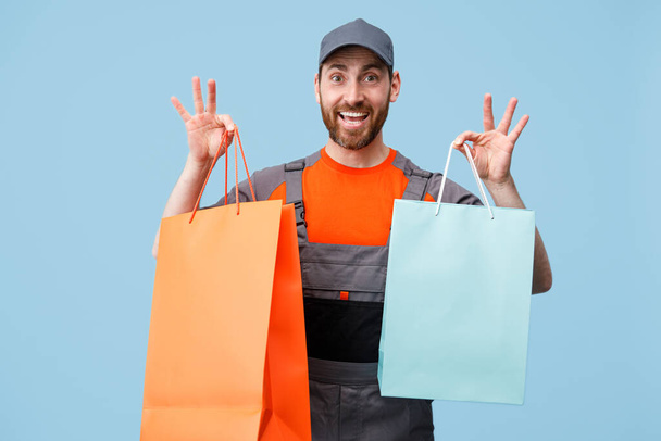 Mensajero masculino sonriente en uniforme sosteniendo bolsas de compras sobre fondo azul. Sesión de estudio - Foto, imagen