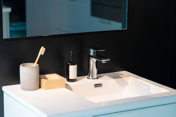 minimale toiletartikelen zoals tandenborstel, serumfles en houtopslag op witte gootsteen en spiegel op de zwarte wand. - Foto, afbeelding