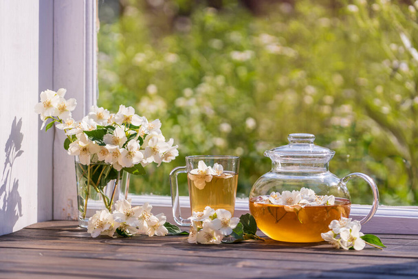 Pyszna gorąca herbata ziołowa na parapecie w domu w letni dzień w pobliżu ogrodu i piękny bukiet kwiatów jaśminu, zbliżenie. Gorąca herbata jaśminowa w szklanym czajniku i filiżance - Zdjęcie, obraz