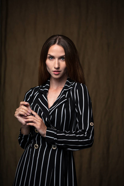 Young woman in black and white striped dress looking at camera posing in studio with dark background - Fotó, kép