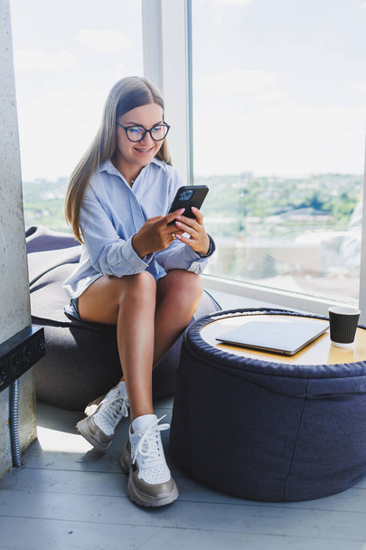 Glückliche Freelancerin mit klassischer Brille, die im modernen Coworking Space am Telefon sitzt und lächelt, unbeschwerte Millennial-Frau mit Brille, die Freizeit zur Kommunikation genießt - Foto, Bild