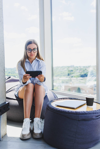 Boldog szabadúszó lány klasszikus szemüvegben nézi a telefont és mosolyog, miközben ül a modern coworking térben, gondtalan Y-generációs nő szemüvegben élvezi a szabadidő a kommunikáció - Fotó, kép
