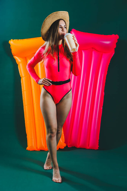 A young woman in a bright pink swimsuit and hat with an inflatable pink mattress on a green background. Summer rest. Girl with a cocktail in her hands in a bathing suit - Foto, Imagen
