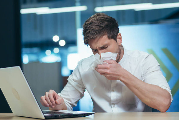 A businessman at work is sick, coughs and has a runny nose, sits at a table with a napkin and sneezes - Photo, image