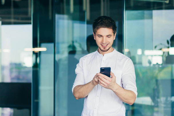 Young businessman standing in the office by the window, looking at the screen of a mobile phone smiling reads an online message - Photo, Image