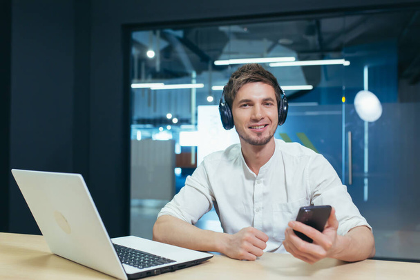 Joven hombre de negocios trabaja en la oficina en un ordenador portátil, escucha música en auriculares grandes utiliza la aplicación para escuchar libros en línea y podcasts utiliza el teléfono - Foto, Imagen