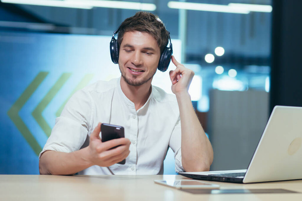 Young businessman works in the office on a laptop, listens to music in big headphones uses the application to listen to online books and podcasts uses the phone - Zdjęcie, obraz