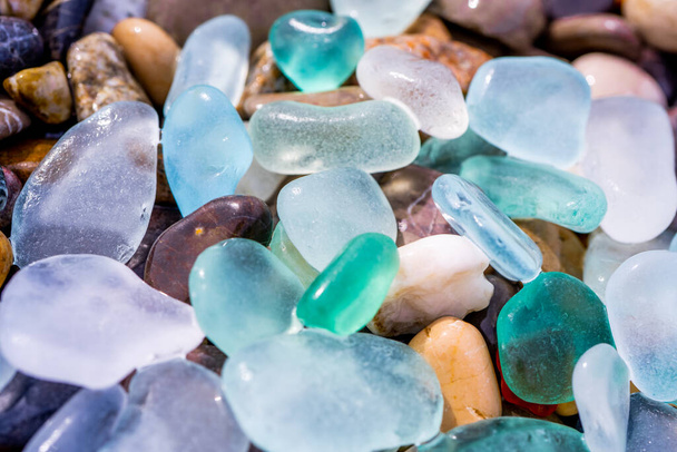 Natural polish textured sea glass and stones on the seashore. Azure clear sea water with waves. Green, blue shiny glass with multi-colored sea pebbles close-up. Beach summer background - Photo, Image