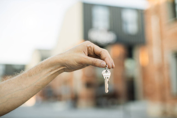 House keys in male hand on background of new buildings and sky. Real estate agent, moving home or renting property. Man hand holds the keys to an apartment against the background of buildings. - Фото, зображення