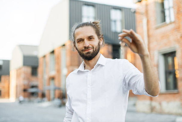 Real estate attractive agent beautiful man with house keys in front of sold apartment, in city street. Young caucasian guy in white shirt with keys just bought new apartment. - Foto, immagini