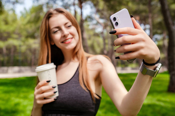 Cute brunette woman wearing sports bra standing on city park, outdoors taking selfie and holding takeaway coffee mug. Self portrait for social media. Outdoor sport concepts. - Zdjęcie, obraz