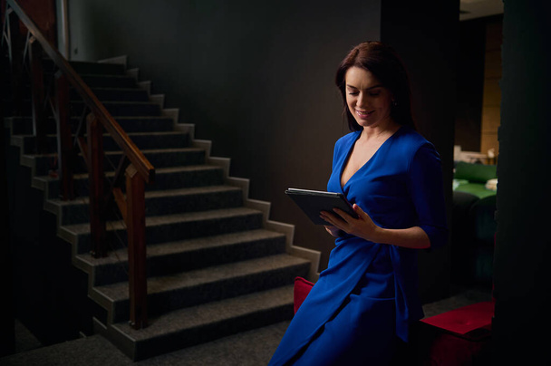 Elegantly dressed attractive dark-haired Caucasian middle-aged woman, sales manager, interior designer working on digital tablet in a stylish interior of home design studio, lit with soft dimmed light - Zdjęcie, obraz