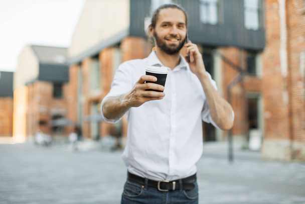 Focus on coffee. Handsome business man holding cup of takeaway and talking on mobile phone. Modern city life in background. Man in white shirt walking and talking on mobile phone. - Фото, зображення