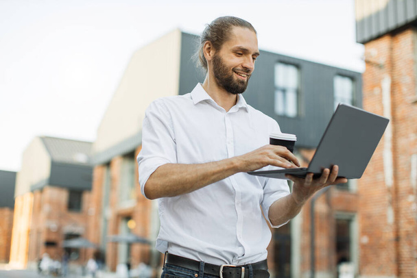 Jovem empresário barbudo atraente em camisa branca trabalhando no laptop. Empresário profissional usando dispositivo digital moderno sorrindo beber café perto de prédio de escritórios na cidade. - Foto, Imagem