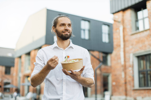 Happy business man eating healthy salad on a break standing over city street background. Male dieting nutrition concept. Attractive smiling guy enjoying veggie meal. - Photo, image