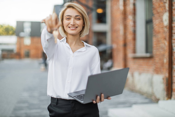 Cheerful young businesswoman in white shirt and black pants holding keys of new flat and laptop, looking at camera and smiling, standing near the building in the middle of the street. - Foto, Imagem