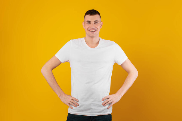 Gelukkige Millennial Man Poseren Handen op Heupen glimlachend naar de camera staan in de studio op gele achtergrond. Shot of Positive Guy draagt een wit T-shirt. Mannelijk modeconcept - Foto, afbeelding