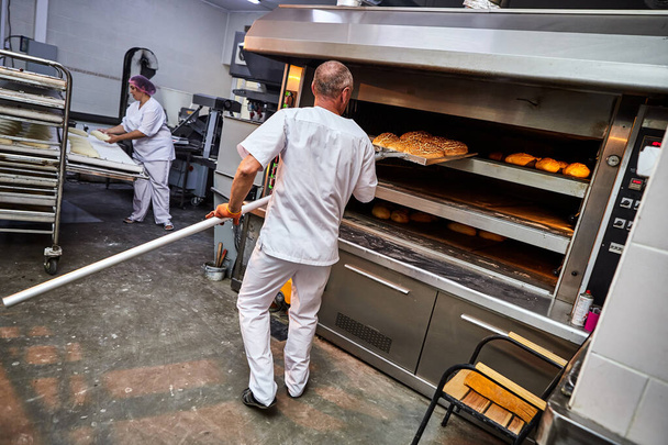 Profesionální pekař v uniformě vytáhne vozík s čerstvě upečeným chlebem z průmyslové pece v pekárně - Fotografie, Obrázek