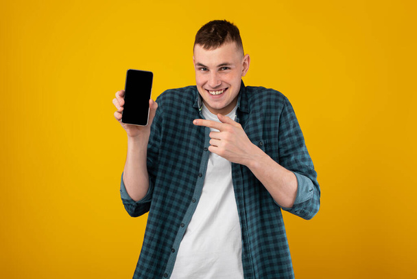 Funny Guy megjelenítése Mobiltelefon mutató ujjal az üres képernyőn Reklám alkalmazás Mosolyogva a kamera felett álló sárga háttér. Remek alkalmazás az okostelefonodhoz. Mockup, Stúdió lövés - Fotó, kép