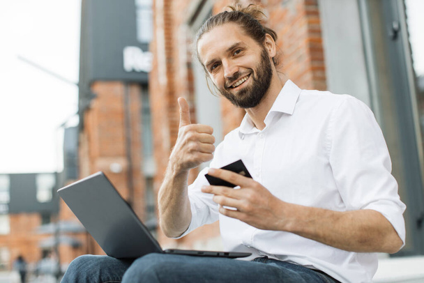 Stylish bearded man doing online purchases while sitting outside near office building. Caucasian guy in formal wear using credit card and laptop for shopping showing thumb up. - Foto, Imagem