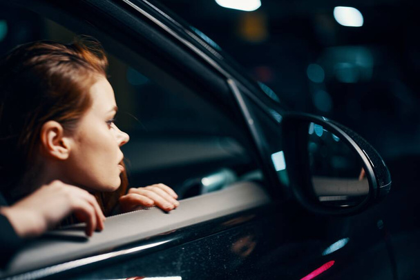 una foto orizzontale ravvicinata dal lato, di notte, di una donna seduta in una macchina nera e che guarda fuori dalla finestra guardando nello specchio laterale. Foto di alta qualità - Foto, immagini