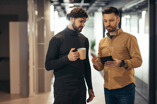 Teamwork, collaboration and coworking concept. Male coworkers discussing business startup project, using digital tablet while walking in company hall during coffee break at modern office - Photo, Image