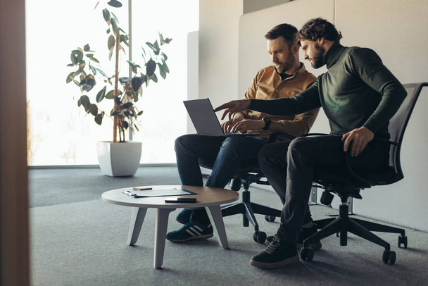 男性の同僚は、ビジネスプロジェクトについて話し合い、アイデアのブレインストーミング、現代のオフィス、空白のスペースで一緒にラップトップコンピュータを使用しています。企業ミーティング、チームワーク、協力の概念 - 写真・画像