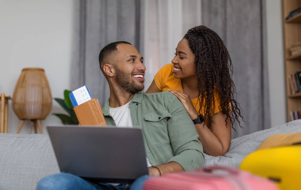 Boldog szerető fekete házastársak készen állnak az utazásra, a beszállókártya tartására és a laptop használatára, együtt tervezik a nyaralást, egymásra néznek és mosolyognak. Technológia és szállítási koncepció - Fotó, kép