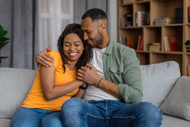 Χαριτωμένοι μαύροι σύζυγοι κάθονται στον καναπέ, αγκαλιάζοντας και χαμογελώντας, απολαμβάνοντας το χρόνο μαζί στο σπίτι. Αφροαμερικανοί σύζυγοι χαλαρώνουν στον καναπέ μαζί. - Φωτογραφία, εικόνα