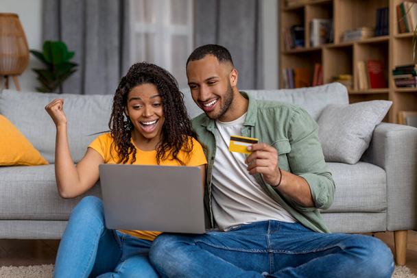 Ja, riesige Umsätze im Webstore. Glückliches schwarzes Paar, das online einkauft, Kreditkarte hält und zu Hause Laptop benutzt. Afroamerikanerin macht Siegergeste mit geballter Faust - Foto, Bild