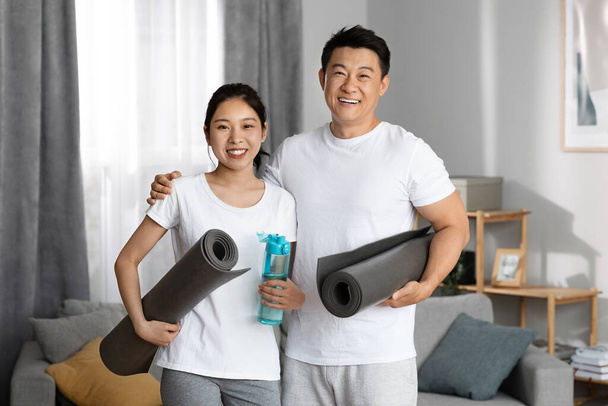 Позитивная азиатская пара собирается в спортзал вместе, счастливый корейский мужчина и женщина в спортивной одежде держа фитнес коврики и бутылку воды, упражнения на дому, имея утреннюю тренировку, копировальное пространство - Фото, изображение
