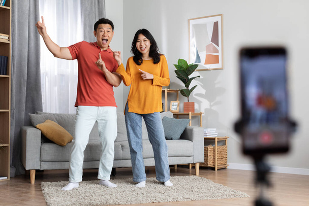 Emotional asian man and woman in casual outfits singing and dancing, having fun and streaming from home, using cell phone set on tripod, positive korean spouses recording video for friends - Photo, Image