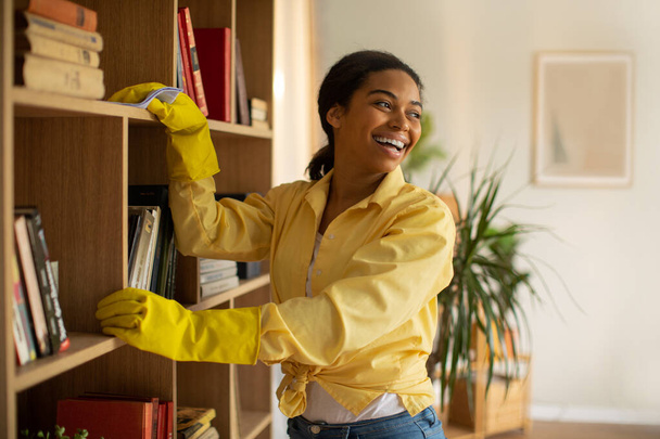 Happy Black Woman Dusting Books Shelves Cleaning Furniture With Detergent And Rag Wearing Yellow Rubber Gloves Standing In Modern Living Room At Home, Smiling Looking Aside. Housework Concept - Foto, immagini