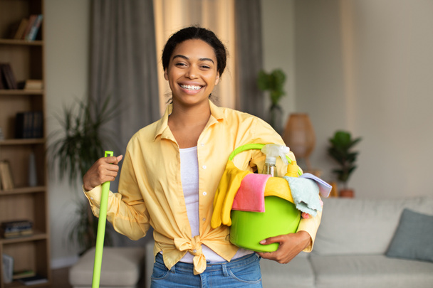 Sprzątanie domu. Szczęśliwa czarna kobieta trzymająca mopa i wiadro z szmatami i detergentami w sprayach i butelkach, udająca uśmiechniętą do kamery stojącą w nowoczesnym salonie w domu. Koncepcja prac domowych - Zdjęcie, obraz