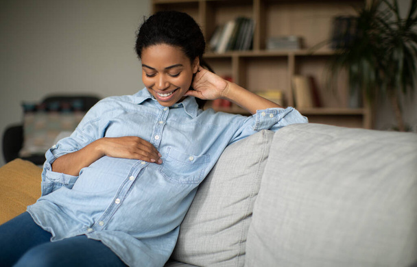 Pregnancy Happiness. Happy Pregnant African American Woman Smiling Stroking Belly Expecting Baby Sitting On Couch At Home. Childbirth And Wellbeing, Maternity Leave Concept - Photo, Image