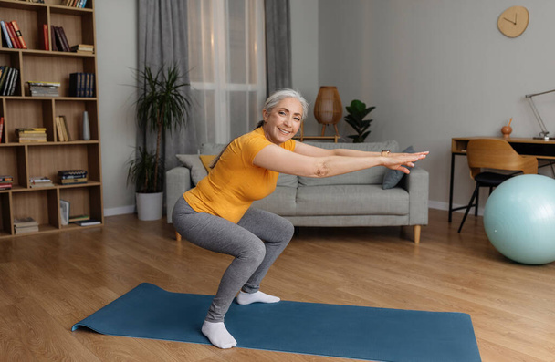 Active senior lady doing squats on domestic workout, training in living room and smiling at camera, copy space. Fit female leading active lifestyle, staying in good shape at home - Photo, Image