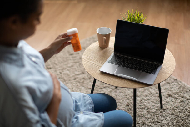Ciężarna Czarna Pani Korzystanie Laptop Holding Medical Pills Bottle Patrząc na pusty ekran komputera o powołanie lekarzy online siedzi w domu. Ciąża, opieka zdrowotna i technologia. Przycięte - Zdjęcie, obraz