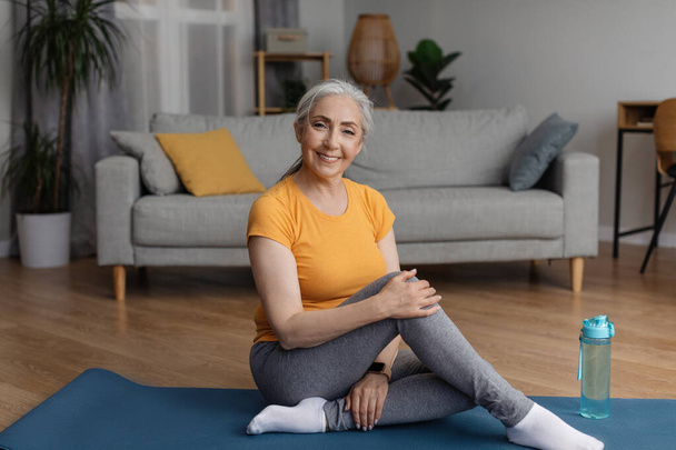 Koncepcja zdrowego stylu życia. Szczęśliwa seniorka siedząca na macie do jogi sportowej w domu w salonie i uśmiechnięta do kamery, kopiująca przestrzeń. Starsza kobieta robi ćwiczenia rehabilitacyjne - Zdjęcie, obraz