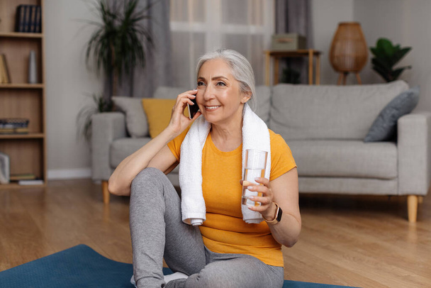 Glückliche Seniorin mit einem Glas Wasser, das in der Pause vom häuslichen Sporttraining auf dem Smartphone spricht, sitzt auf einer Yogamatte. Positive Frauen genießen Telefongespräch nach dem Heimtraining - Foto, Bild