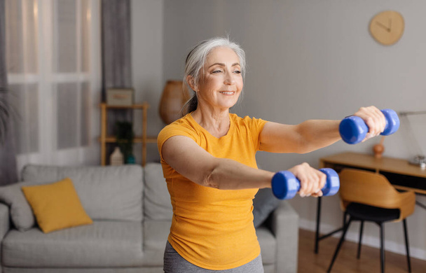 Возбужденная пожилая женщина, занимающаяся домашними тренировками, тренирующаяся с гантелями, упражняющая бицепсы дома, в свободном пространстве. Подходящая пожилая женщина, ведущая активный образ жизни - Фото, изображение