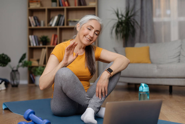 Pozytywna seniorka machająca do laptopa kamerką internetową, robiąca wideokonferencję z osobistym trenerem, siedząca na macie do jogi przed komputerem, gotowa do treningu domowego - Zdjęcie, obraz
