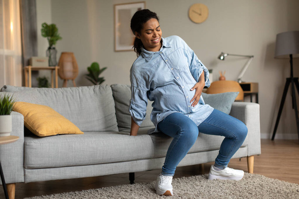 Grávida preto fêmea pé de sofá tocando barriga grande sofrendo de dor tendo contrações espasmo em casa. Conceito de parto, parto e cuidados de saúde - Foto, Imagem
