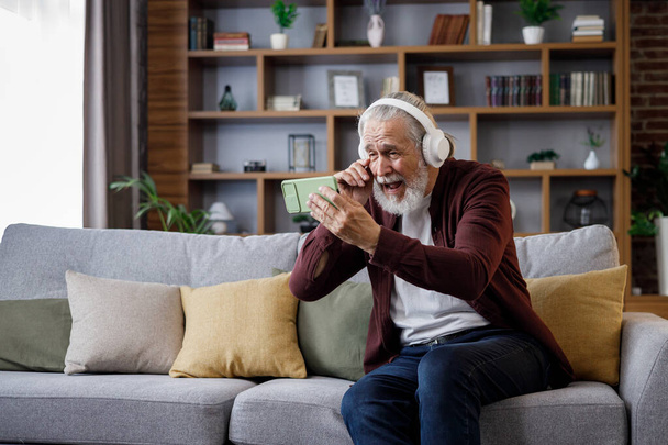 Happy handsome elderly gray-haired man in white headphones holding smartphone and laughing. Old man sitting on couch at home watching funny content videos blogs, TV shows, films. - Foto, imagen